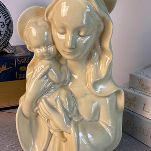Puede incluir: Una estatua de cerámica color crema de María sosteniendo al niño Jesús. La estatua tiene un acabado liso y brillante y está detallada con túnicas fluidas y halos. Los ojos de las figuras están cerrados, transmitiendo una sensación de paz y serenidad. Es un artículo de decoración religiosa.