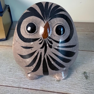 May include: A ceramic owl figurine with a rounded body and small feet. The owl is primarily light brown with black and brown decorative markings. The eyes are large and black, and the beak is brown. The owl is a decorative item.