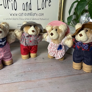 May include: Four plush teddy bears dressed in various outfits. One bear wears a blue bandana and red striped overalls. Another is in a red and black plaid shirt and red pants. A third bear is in a pink dress and hat. The last bear wears a plaid shirt, denim shorts, and a bow tie.