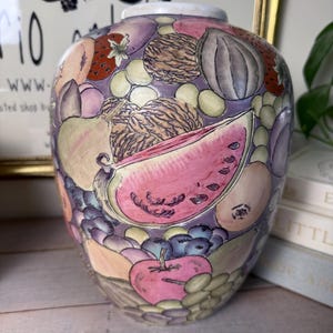 May include: A decorative ceramic vase with a colorful fruit design. The vase features a variety of fruits, including watermelon, apples, grapes, and strawberries, in shades of pink, purple, and green. The vase has a rounded shape and a white rim.