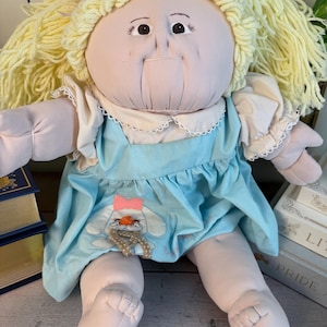 Puede incluir: Muñeca vintage con cuerpo de tela suave y cara redonda. La muñeca tiene cabello de hilo amarillo y viste un vestido azul claro con cuello blanco y un aplique de conejo. Un juguete clásico.