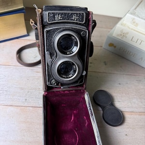 May include: Vintage twin-lens reflex camera in a brown leather case. The camera has two lenses and a black body with silver accents. The case is open, revealing a burgundy velvet interior. The camera has Japanese text on the top.