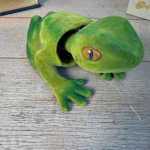 May include: A plush green frog with a large open mouth, brown eyes, and outstretched arms. The frog is made of a fuzzy material and has a detailed design. The frog is a decorative item.