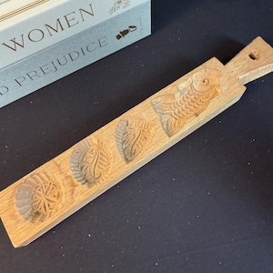 May include: A wooden cookie mould featuring four carved designs: a flower, a leaf, another leaf, and a fish. The mould is rectangular with a handle and a hole for hanging. The wood has a natural, light brown colour.