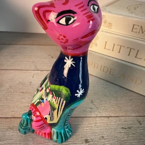 May include: A hand-painted ceramic cat figurine. The cat has a pink head with black and white facial details, a blue collar with a white star, and a colorful body with green, pink, and yellow accents. The cat is sitting on a wooden surface.