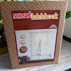 May include: A Sunset Latchhook kit, titled "White Splendor", is displayed in its original packaging. The kit includes materials for a wall hanging with a macrame design, featuring white yarn and a wooden dowel. The box is brown with the brand name repeated.