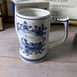 May include: White ceramic mug with a blue and white floral design and a landscape scene. The mug has a rectangular shape with a handle and the word "Heineken" is printed on the front. The mug is approximately 4 inches tall.