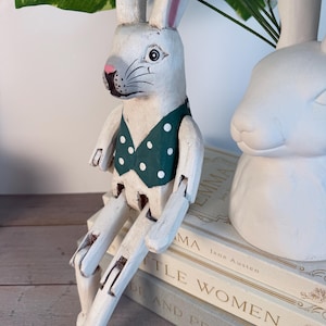 May include: A white wooden rabbit figurine with articulated limbs, wearing a green vest with white polka dots. The rabbit has pink inner ears and a pink nose. It is sitting on a stack of books, with another white rabbit figurine in the background.