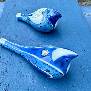 May include: Two ceramic bird figurines in shades of blue, white, and speckled accents. The birds have a glossy finish and are arranged on a blue surface. The design features a mix of solid and patterned areas.