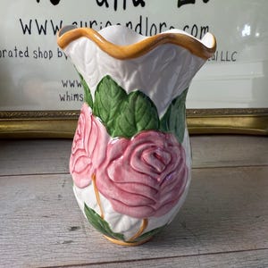 Puede incluir: Jarrón de cerámica blanca con un diseño de rosa rosa. El jarrón tiene una parte superior con volantes con un borde dorado y hojas verdes. El jarrón está sobre una superficie de madera.
