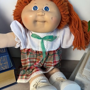 May include: A vintage doll with a round face, blue eyes, and orange yarn hair. The doll wears a white dress with a green ribbon, a plaid skirt, white socks, and white shoes. The doll is sitting on a wooden surface.