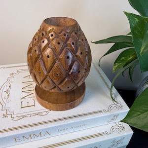 May include: A carved wooden pineapple-shaped decorative item with a round base. The item has a series of diamond-shaped cutouts and a hollowed-out top. It sits atop a stack of books with the title "Emma" in gold lettering.