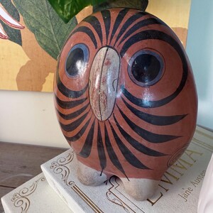 May include: A ceramic owl figurine with a reddish-brown body and black striped design. The owl has large black eyes with blue accents and a white beak. The figurine is perched on top of a stack of books.