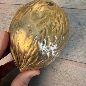 Puede incluir: Un objeto decorativo dorado en forma de huevo con una superficie texturizada. El objeto se sostiene en una mano y el fondo es una superficie de madera.