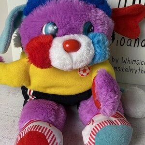 May include: A colorful plush dog toy with a red nose, blue eyes, and a soccer ball emblem. The dog has a purple body, blue and red accents, and wears a yellow shirt. The toy's feet are red and blue with white stripes.