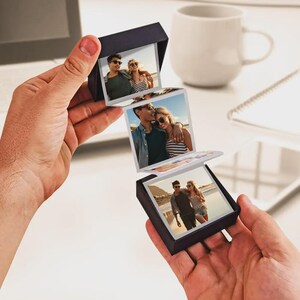 Puede incluir: Una caja de fotos negra en forma de acordeón que muestra tres imágenes de una pareja. La caja está abierta, revelando las fotografías. Las imágenes muestran a la pareja en una playa, con gafas de sol y ropa informal. La caja se sostiene frente a un fondo blanco.