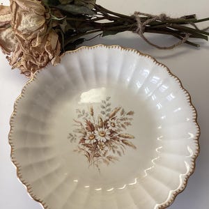 May include: A white ceramic bowl with a gold rim and a floral design featuring brown and white daisies and wheat. The bowl has a scalloped edge and is perfect for serving snacks or desserts.