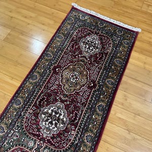 Może przedstawiać: Czerwony, zielony i czarny wzorzysty dywanik z białą frędzlą. Dywanik ma tradycyjny perski wzór.