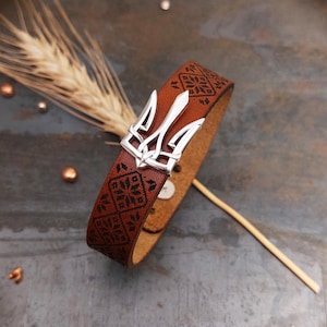 May include: A brown leather bracelet with a silver trident clasp. The bracelet is decorated with a black geometric pattern.