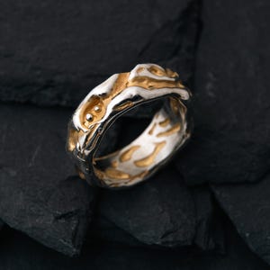 May include: A two-tone ring with a textured design. The ring features a silver band with gold-colored accents. The top of the ring has a raised, organic shape with two small, round gold details. The ring is set against a dark, textured background.