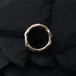 May include: A gold and silver ring with an organic, textured design. The ring has a band of gold and a silver-colored, irregular outer edge. The ring is set against a dark, textured background.
