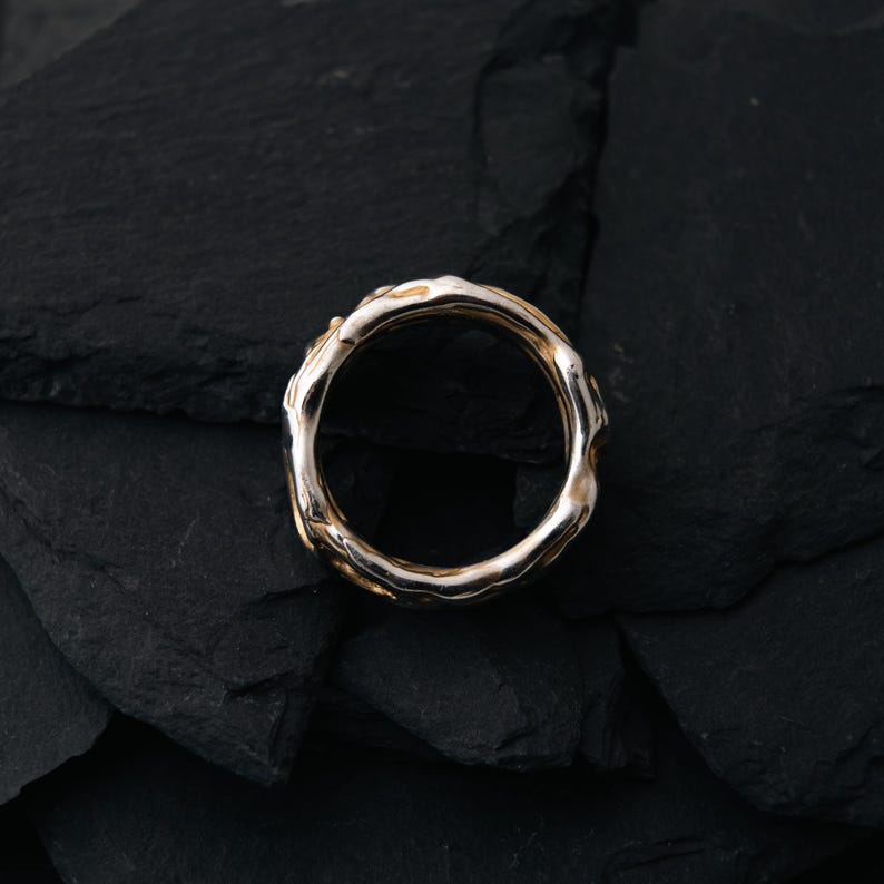 May include: A gold and silver ring with an organic, textured design. The ring has a band of gold and a silver-colored, irregular outer edge. The ring is set against a dark, textured background.