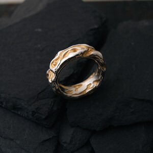 May include: A two-tone ring with a textured design. The ring features a silver band with gold-colored accents. The design has an organic, flowing pattern. The ring is displayed on a dark, textured surface.