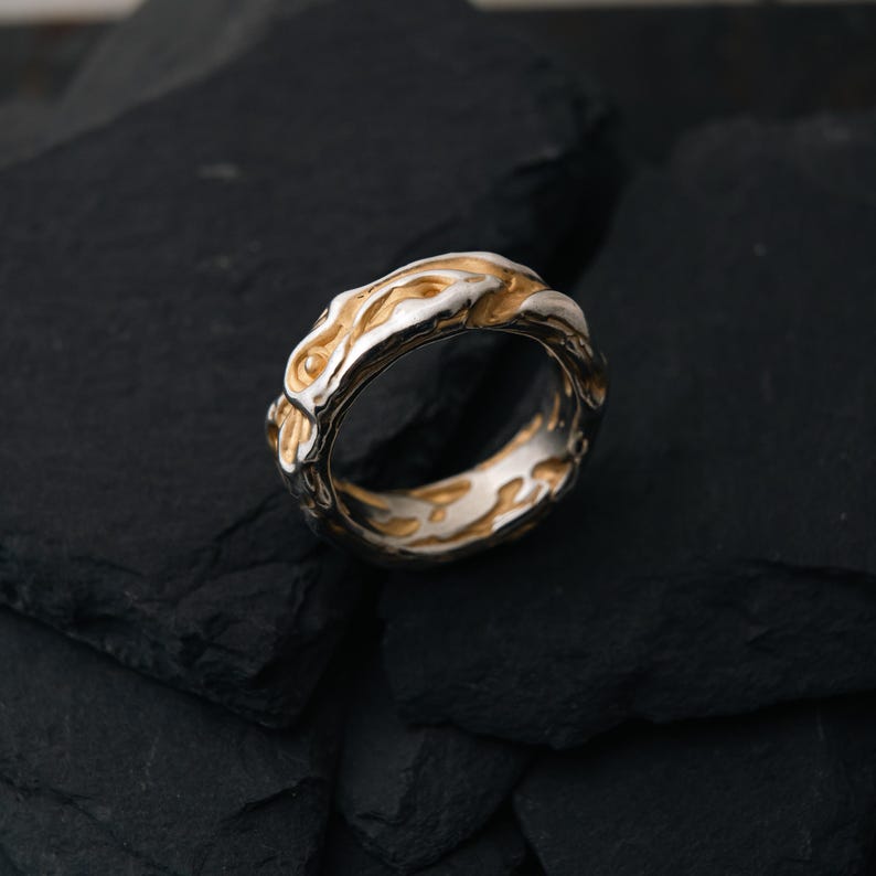 May include: A two-tone ring with a textured design. The ring features a silver band with gold-colored accents. The design has an organic, flowing pattern. The ring is displayed on a dark, textured surface.
