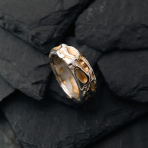 May include: A silver ring with a textured surface and gold-colored accents. The ring has an organic, irregular pattern, and is set against a dark, textured background. The ring's design is modern and unique.