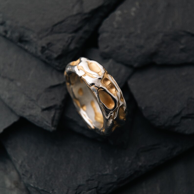 May include: A silver ring with a textured surface and gold-colored accents. The ring has an organic, irregular pattern, and is set against a dark, textured background. The ring's design is modern and unique.