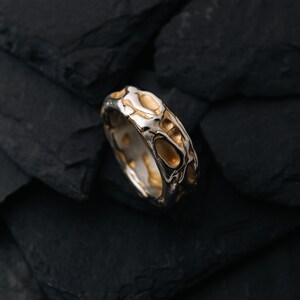 May include: A silver ring with a textured design featuring gold-colored accents. The ring has an organic, irregular pattern, with a mix of polished and textured surfaces. The ring is set against a dark, textured background.