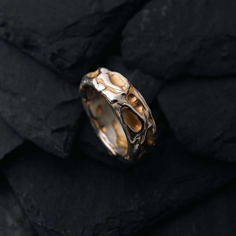 May include: A silver ring with a textured design featuring gold-colored accents. The ring has an organic, irregular pattern, with a mix of polished and textured surfaces. The ring is set against a dark, textured background.