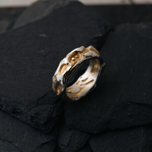 May include: A two-tone ring with a textured, organic design. The ring features a silver band with gold-colored accents, creating a striking contrast. The design has an irregular, flowing shape, adding to its unique appeal. The ring is likely made of metal.