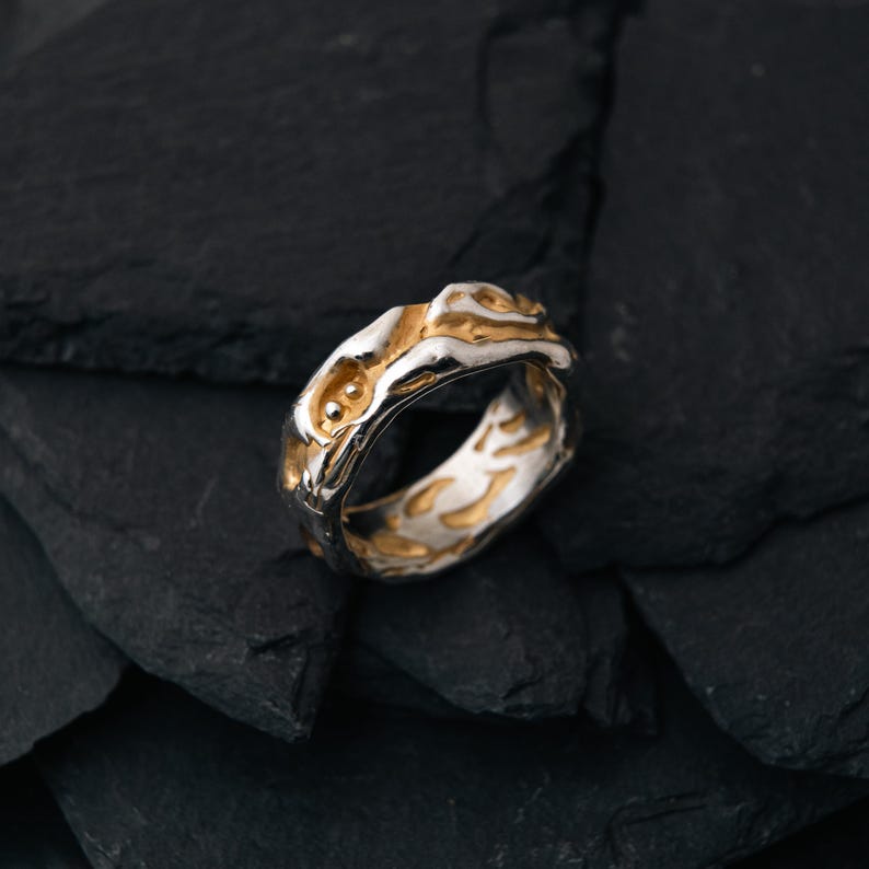 May include: A two-tone ring with a textured design. The ring features a silver band with gold-colored accents. The top of the ring has a raised, organic shape with two small, round gold details. The ring is set against a dark, textured background.