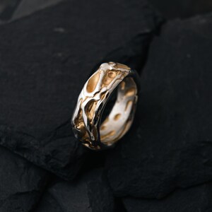 May include: A two-tone ring with a textured, organic design. The ring features a silver band with gold-colored accents, creating a striking contrast. The surface has an irregular, almost molten appearance, adding to its unique aesthetic.