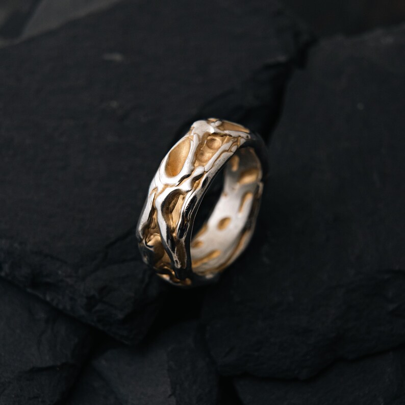 May include: A two-tone ring with a textured, organic design. The ring features a silver band with gold-colored accents, creating a striking contrast. The surface has an irregular, almost molten appearance, adding to its unique aesthetic.