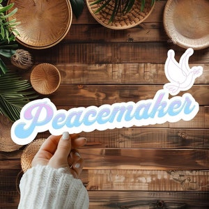 May include: A sticker with a blue and pink gradient design that says "Peacemaker" in a stylized font. A white outline of a dove with an olive branch in its beak is above the word.