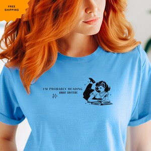 May include: A light blue t-shirt with the text "I'M PROBABLY READING ABOUT SHIFTERS" and a black and white illustration of a woman reading a book. The shirt has short sleeves and a crew neck.