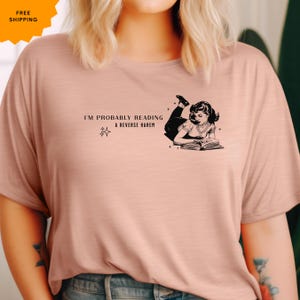 Könnte beinhalten: Ein rosa T-Shirt mit dem Text "I'm probably reading a reverse harem" und einer Schwarzweißillustration einer Frau, die ein Buch liest.