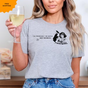 Peut inclure: T-shirt gris avec une illustration en noir et blanc d'une femme lisant un livre. Le texte sur le t-shirt est "Je lis probablement des histoires de loups-garous."