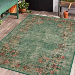 May include: A rectangular green rug with a floral pattern in shades of pink, beige, and cream. The rug has a distressed look and a dark green border. The rug is placed on a wooden floor, with a green bench and plants in the background.