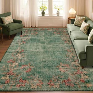 May include: A large, rectangular, distressed-look rug with a teal background and a floral border in shades of pink, cream, and green. The rug is placed in a living room setting with green furniture and natural light.