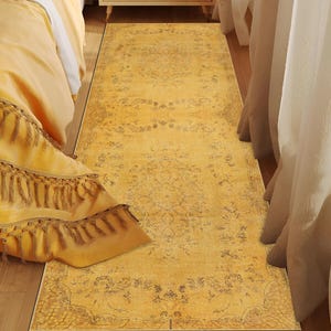 Può includere: Un lungo tappeto rettangolare giallo con un motivo floreale sbiadito. Il tappeto è posizionato su un pavimento in legno accanto a un letto con biancheria da letto gialla e un comodino. Il design del tappeto include un bordo e un medaglione centrale. Misura circa 200 cm di lunghezza.
