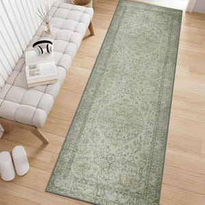 May include: A long rectangular runner rug featuring a faded sage green and cream color palette with an intricate, symmetrical floral medallion pattern and a decorative border, laid flat on a light-colored wooden floor surface.