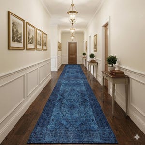 May include: A long, ornate blue runner rug with a detailed floral pattern, placed in a hallway with dark wood flooring. The walls are cream-colored with white wainscoting and framed artwork. Overhead, there are decorative hanging lights.