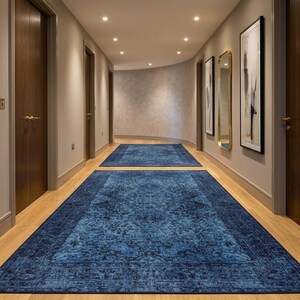 May include: A hallway with two blue patterned rugs. The rugs are rectangular and have a dark blue border. The walls are light beige, and the doors are dark brown. Artwork and a mirror hang on the wall.