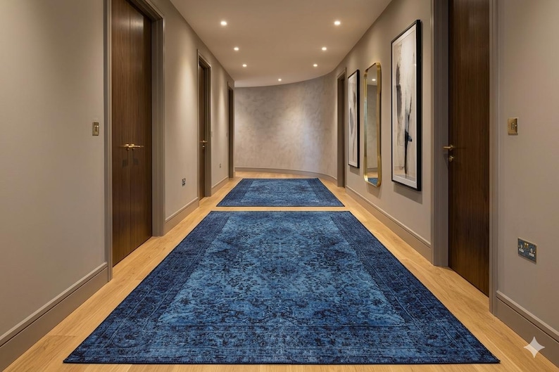 May include: A hallway with two blue patterned rugs. The rugs are rectangular and have a dark blue border. The walls are light beige, and the doors are dark brown. Artwork and a mirror hang on the wall.
