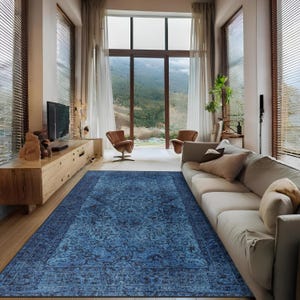 May include: A large, rectangular blue rug with an intricate floral pattern dominates the living room. The rug is placed on a wooden floor, and the room features a long, light-colored sofa, two brown chairs, and a wooden entertainment center. Large windows offer a view of a mountain landscape.