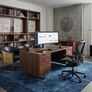 May include: A modern home office with a large wooden desk, a black ergonomic chair, and a blue patterned rug. A large computer monitor displays data. Bookshelves and a blue sofa are also visible.