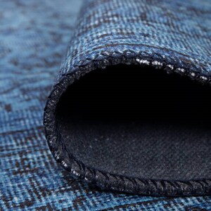 May include: Close-up of a rolled-up blue rug with a textured pattern. The rug's edge reveals a dark blue backing and a stitched border. The rug's surface has a faded, distressed appearance, with shades of blue and black.